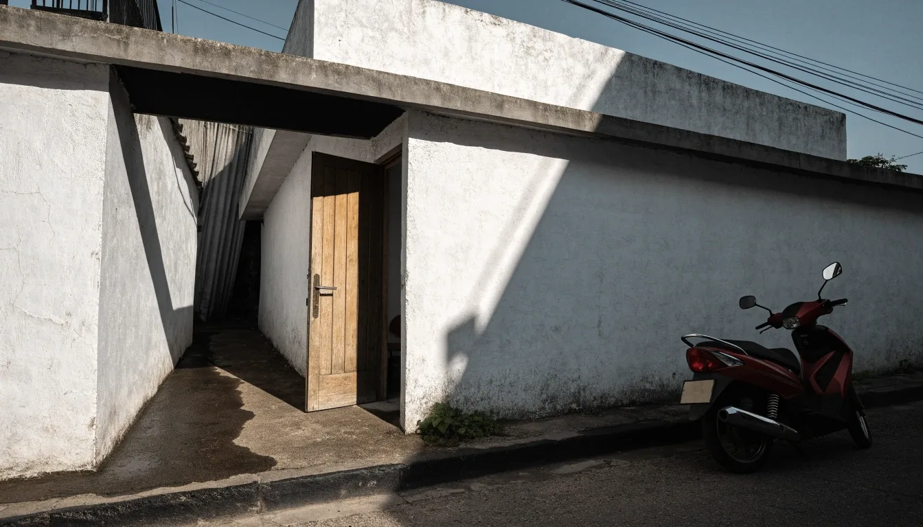 Low-rise administrative clinic building with a whitewashed wall, deep awning shadow, and an ajar wooden door seen from the street at noon